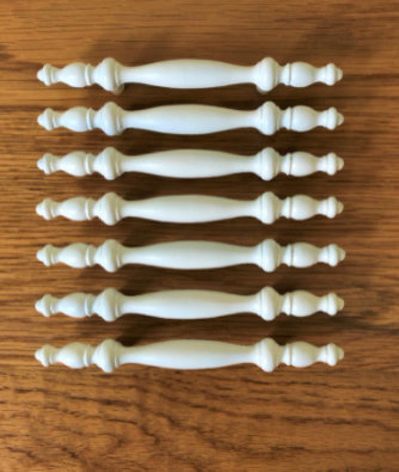 May include: Set of seven white cabinet knobs with a decorative, slightly curved design. The knobs have a smooth, matte finish.
