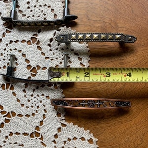 May include: Four different cabinet door pulls. Two are black with gold accents, one is copper with a floral design, and one is black with gold pyramid-shaped accents.