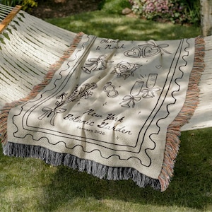 May include: A cream-colored throw blanket with black line art illustrations of wedding-themed icons, including bells, hearts, and champagne glasses. The blanket has a decorative border and the text "Olivia & Noah" and "New York Botanic Garden January 2026".