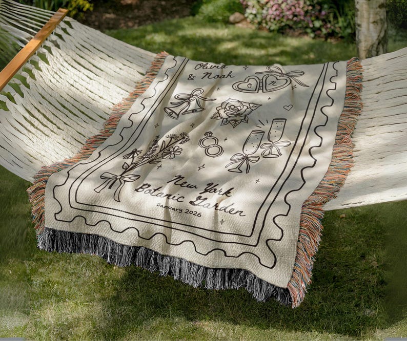 May include: A cream-colored throw blanket with black line art illustrations of wedding-themed icons, including bells, hearts, and champagne glasses. The blanket has a decorative border and the text "Olivia & Noah" and "New York Botanic Garden January 2026".