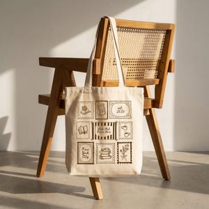 May include: A beige canvas tote bag featuring a brown graphic print of nine postage stamp-style illustrations. The central stamp displays the text "Book Club Name Here" surrounded by stripes, with other stamps showing books, flowers, and "est. 2025".