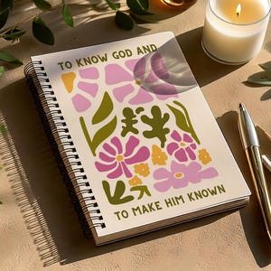 Op de afbeelding: Spiraalgebonden notitieboek met de tekst "TO KNOW GOD AND TO MAKE HIM KNOWN" en een bloemenontwerp in roze, geel en groen. Een gouden pen en een brandende kaars liggen naast het notitieboek.