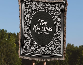 Könnte beinhalten: Eine schwarze Webdecke mit weißen Linienzeichnungen, die einen zentralen ovalen Rahmen mit dem Text The Kellums EST. 2026 enthält. Der Rand ist mit Blumenmustern verziert und die Kanten der Decke weisen an allen Seiten eine Fransenstruktur auf.