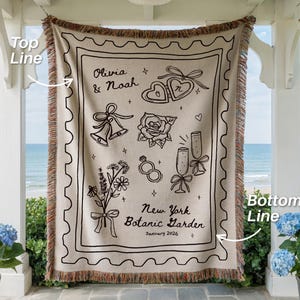 May include: Beige throw blanket with black line art of wedding-themed illustrations, including hearts, bells, and flowers. The text reads "Olivia & Noah" and "New York Botanic Garden January 2026". The blanket has a fringed edge.