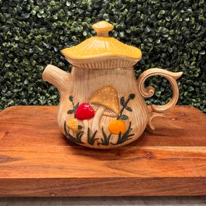 Peut inclure: Une théière en céramique en forme de champignon avec un corps beige à texture de grain de bois et un couvercle jaune à pois. L'avant présente des illustrations de champignons en relief rouge, orange et jaune, avec de l'herbe et des tiges vertes.