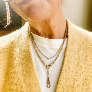 Pode incluir: Um colar de corrente de ouro com um pequeno pingente e uma corrente mais longa com um fecho decorativo. O colar &eacute; usado sobre uma camisa branca e um cardigan amarelo.