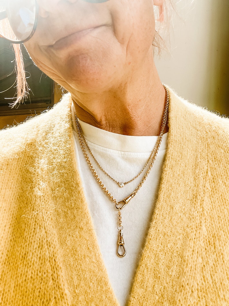 Pode incluir: Um colar de corrente de ouro com um pequeno pingente e uma corrente mais longa com um fecho decorativo. O colar &eacute; usado sobre uma camisa branca e um cardigan amarelo.