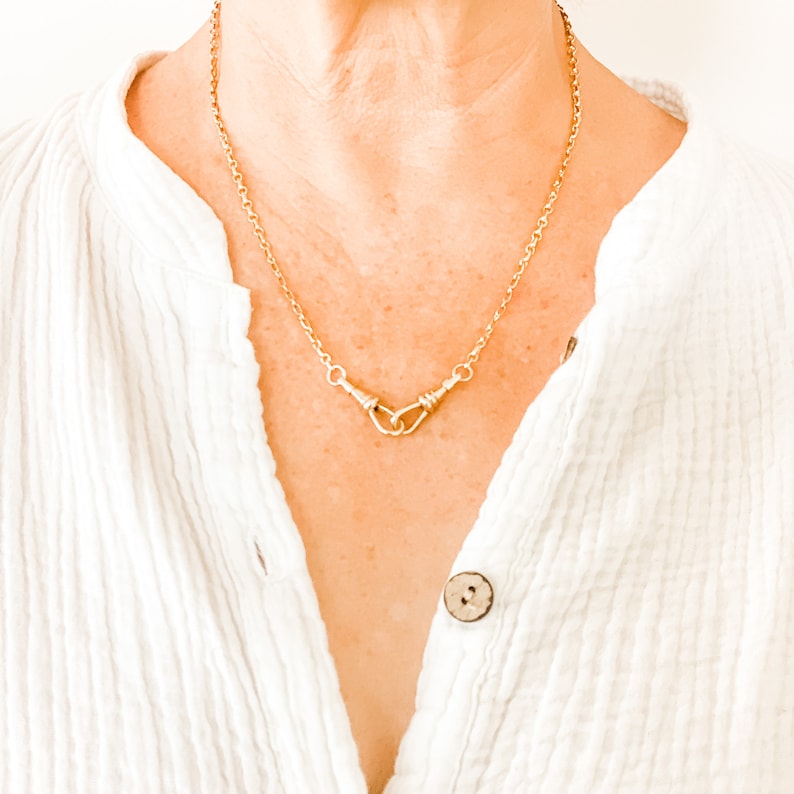 May include: A gold chain necklace with a unique double-loop pendant. The necklace is worn by a person with a white shirt.