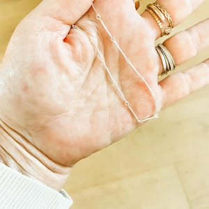 Peut inclure: Un délicat collier en argent avec un design linéaire. Le collier présente de petits maillons rectangulaires reliés par de fins anneaux circulaires. Le collier est tenu dans une main avec plusieurs bagues en or et en argent.
