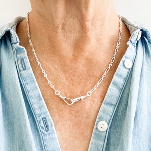 Puede incluir: Un collar de cadena de plata con un diseño de cierre único. La cadena está hecha de pequeños óvalos unidos. El cierre es un diseño de doble gancho.