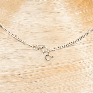 Peut inclure: Un collier chaîne gourmette argenté avec une fermeture à fermoir mousqueton. Le collier est présenté sur une surface en bois brun clair. Les maillons de la chaîne sont étroitement liés.