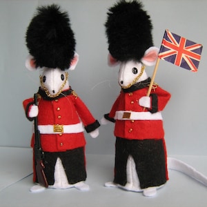Pode incluir: Duas figurinhas de rato branco vestidas como guardas britânicos com uniformes vermelhos e pretos e chapéus de pele preta. Um rato segura um rifle e o outro uma pequena bandeira da Union Jack.