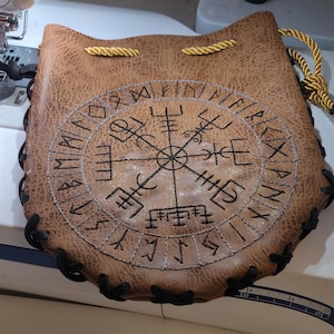May include: Brown leather pouch with a drawstring closure and a stitched black border. The front features a detailed embroidered design of a compass rose with runic symbols in black thread. A gold cord is used for the drawstring.