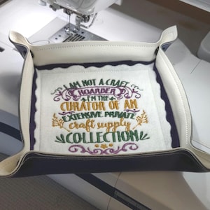 May include: A square, two-tone leather tray with a white interior and dark blue exterior. The tray features the embroidered text: "I AM NOT A CRAFT HOARDER I'M THE CURATOR OF AN EXTENSIVE PRIVATE craft supply COLLECTION."