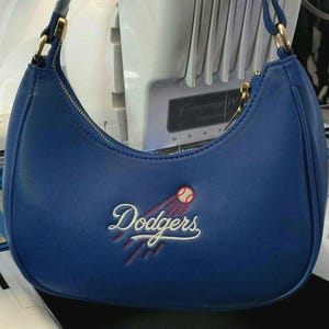 May include: A blue shoulder bag with a curved shape. The bag features a gold zipper and strap hardware. The word "Dodgers" is embroidered in white script with a baseball graphic. The bag is made of a smooth material.