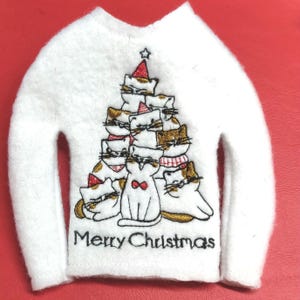 May include: White felt Christmas sweater with a cat-shaped tree design. The tree is made of embroidered cats wearing Santa hats and scarves. The words "Merry Christmas" are embroidered in black at the bottom.