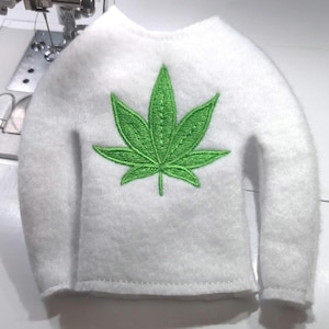 May include: A white felt sweater with a green embroidered cannabis leaf design. The sweater has long sleeves and a crew neck. The cannabis leaf is centered on the front of the sweater.