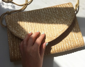 May include: A light brown woven straw clutch bag with a flap closure. The bag has a woven texture and a light brown strap.