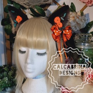Puede incluir: Un par de orejas de gato de piel sintética marrón con calabazas de fieltro naranja y hojas de fieltro verde. Las orejas están unidas a una diadema negra con un lazo de lunares rojos y blancos y una cinta naranja.