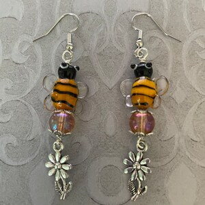 May include: A pair of silver dangle earrings featuring a glass bee charm with black and yellow stripes, a iridescent purple bead, and a silver daisy charm.