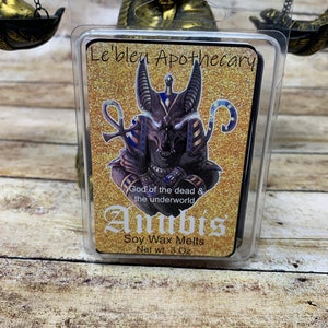 May include: A clear plastic package containing a black soy wax melt with a gold glitter background. The wax melt features an image of Anubis, the Egyptian god of the dead, with the text "Le'bleu Apothecary", "God of the dead & the underworld", "Anubis", "Soy Wax Melts", and "Net wt. 3 Oz".