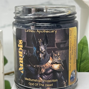 May include: A black glass candle jar with a gold label featuring an image of Anubis, the Egyptian god of the dead. The label also includes the text "Mediumship Protection God of The Dead" and "Le'bleu Apothecary". The candle has notes of cypress, ambergris, and dried tobacco.
