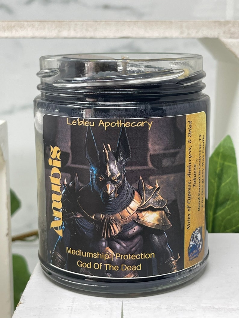 May include: A black glass candle jar with a gold label featuring an image of Anubis, the Egyptian god of the dead. The label also includes the text "Mediumship Protection God of The Dead" and "Le'bleu Apothecary". The candle has notes of cypress, ambergris, and dried tobacco.