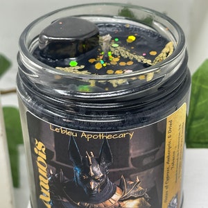 May include: Black candle jar with a gold lid and a black and gold label. The label features an image of Anubis, the Egyptian god of the dead, and the text "Anubis, God of the Dead, Mediumship, Protection, Le'bleu Apothecary, Notes of Cypress, Ambergris, & Dried Tobacco".