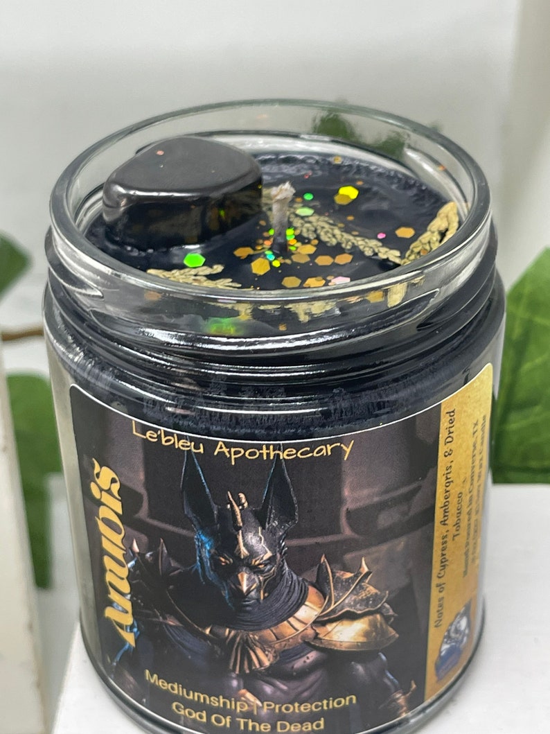 May include: Black candle jar with a gold lid and a black and gold label. The label features an image of Anubis, the Egyptian god of the dead, and the text "Anubis, God of the Dead, Mediumship, Protection, Le'bleu Apothecary, Notes of Cypress, Ambergris, & Dried Tobacco".