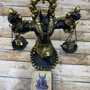 May include: A black and gold Anubis statue with a skull-shaped halo. The statue is holding scales with a heart and a snake on them. The statue is on a wooden surface. The image also shows a soy wax melt with a picture of Anubis on the front. The wax melt is labeled "Le'bleu Apothecary" and "Anubis God of the dead & the underworld Soy Wax Melts Net Wt. 3 Oz."