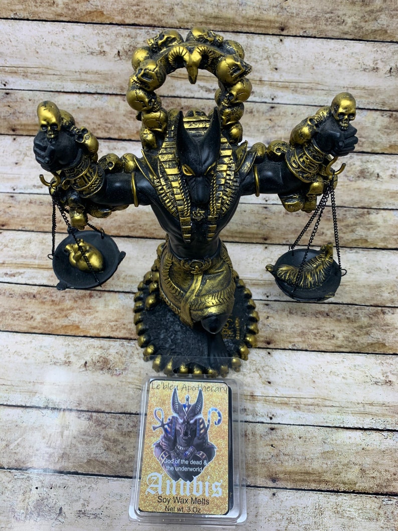 May include: A black and gold Anubis statue with a skull-shaped halo. The statue is holding scales with a heart and a snake on them. The statue is on a wooden surface. The image also shows a soy wax melt with a picture of Anubis on the front. The wax melt is labeled "Le'bleu Apothecary" and "Anubis God of the dead & the underworld Soy Wax Melts Net Wt. 3 Oz."