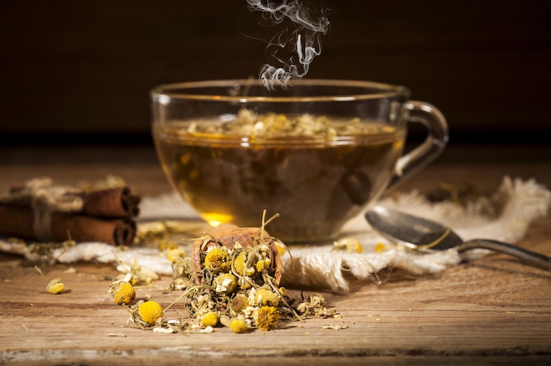 May include: A steaming cup of chamomile tea with dried chamomile flowers scattered on a wooden table.