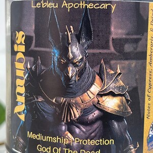 May include: Black and gold candle jar with an image of Anubis, the Egyptian god of the dead. The label reads "Anubis Mediumship | Protection God Of The Dead" and "Le'bleu Apothecary". Notes of Cypress, Ambergris, & Priad.