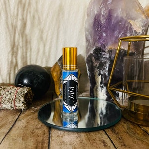 Pode incluir: Um pequeno frasco de vidro com uma tampa dourada e um rótulo azul e branco que diz "Le Petit Apothecary" e "Isis" em forma de diamante. O frasco está sobre um pequeno espelho redondo.