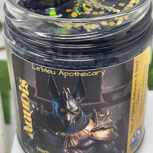 May include: Black candle jar with a gold glitter top. The jar features a black and gold image of Anubis, the Egyptian god of the dead. The text "Le'bleu Apothecary" is printed on the jar. The text "Mediumship Protection God Of The Dead" is printed below the image. The text "Notes of Cypress, Ambergris, & Dried Tobacco" is printed on the right side of the jar.