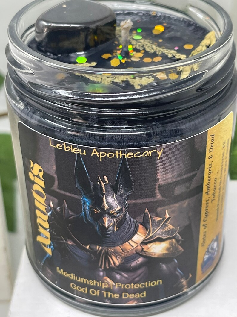 May include: Black candle jar with a gold glitter top. The jar features a black and gold image of Anubis, the Egyptian god of the dead. The text "Le'bleu Apothecary" is printed on the jar. The text "Mediumship Protection God Of The Dead" is printed below the image. The text "Notes of Cypress, Ambergris, & Dried Tobacco" is printed on the right side of the jar.