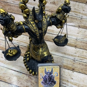 May include: A black and gold Anubis statue with a skull-shaped crown and scales. The statue is holding a black and gold scale in each hand. A small package of soy wax melts with an image of Anubis and the text "Le'bleu Apothecary God of the dead & the underworld Anubis Soy Wax Melts Net wt. 3 Oz" is in the foreground.