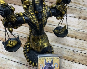 May include: A black and gold Anubis statue with a skull-shaped crown and scales. The statue is holding a black and gold scale in each hand. A small package of soy wax melts with an image of Anubis and the text "Le'bleu Apothecary God of the dead & the underworld Anubis Soy Wax Melts Net wt. 3 Oz" is in the foreground.