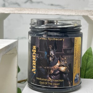 May include: Black candle jar with a gold label featuring an illustration of Anubis, the Egyptian god of the dead. The label reads "Anubis, God of the Dead, Mediumship Protection, Le'bleu Apothecary, Notes of Cypress, Ambergris, & Dried Tobacco, Hand Poured in Converse, TX, 9 oz (227g) Soy Wax Candle."