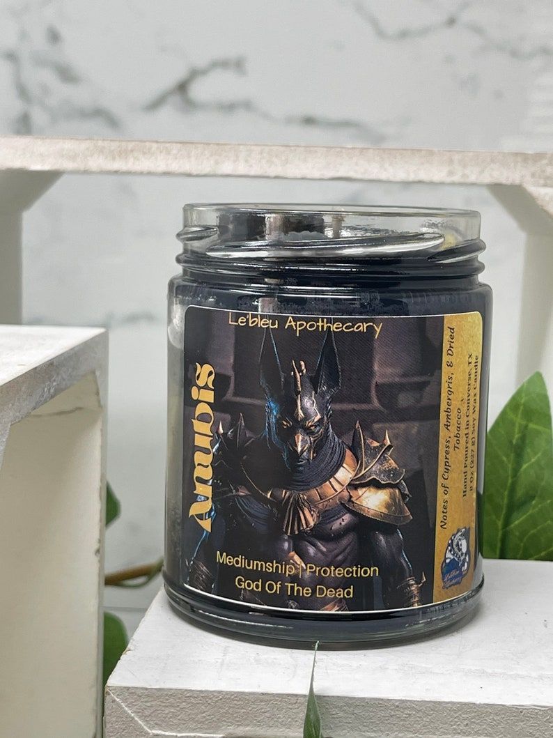 May include: Black candle jar with a gold label featuring an illustration of Anubis, the Egyptian god of the dead. The label reads "Anubis, God of the Dead, Mediumship Protection, Le'bleu Apothecary, Notes of Cypress, Ambergris, & Dried Tobacco, Hand Poured in Converse, TX, 9 oz (227g) Soy Wax Candle."