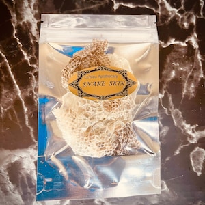 Puede incluir: Una bolsa de plástico transparente que contiene una piel de serpiente seca. La bolsa tiene una etiqueta dorada que dice "Le'bleu Apothecary Snake Skin".
