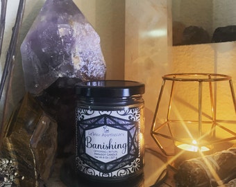 May include: A glass jar candle with a black lid and a label that reads "Banishing" is centered on a mirrored surface. The candle is surrounded by crystals, a gold geometric candle holder, and other decorative items. The candle weighs 227 g.