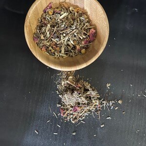 May include: A wooden bowl filled with a blend of dried herbs and flowers, including lavender, chamomile, and rose petals. Some of the herbs and flowers have spilled out of the bowl onto a black surface.