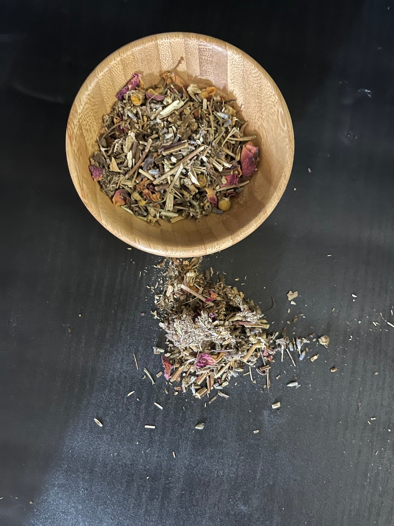 May include: A wooden bowl filled with a blend of dried herbs and flowers, including lavender, chamomile, and rose petals. Some of the herbs and flowers have spilled out of the bowl onto a black surface.