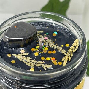 May include: A black candle with gold glitter and dried botanicals in a glass jar. The candle has a black stone on top and the label "Lebleu Apothecary" is visible on the bottom of the jar.