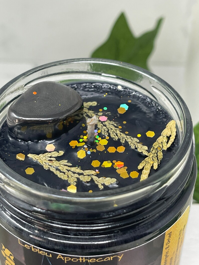 May include: A black candle with gold glitter and dried botanicals in a glass jar. The candle has a black stone on top and the label "Lebleu Apothecary" is visible on the bottom of the jar.