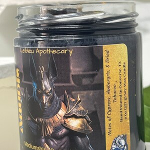 May include: Black glass candle jar with a gold and black label featuring an image of Anubis, the Egyptian god of the dead. The label reads "Lebleu Apothecary", "Mediumship | Protection", "God of the Dead", "Notes of Cypress, Ambergris, & Dried Tobacco", "Hand Poured in Converse, TX", "8 Oz (227 g) Soy Wax Candle".