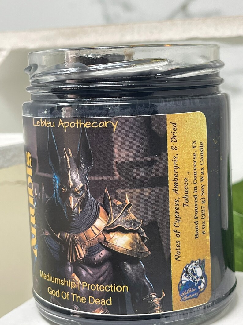 May include: Black glass candle jar with a gold and black label featuring an image of Anubis, the Egyptian god of the dead. The label reads "Lebleu Apothecary", "Mediumship | Protection", "God of the Dead", "Notes of Cypress, Ambergris, & Dried Tobacco", "Hand Poured in Converse, TX", "8 Oz (227 g) Soy Wax Candle".