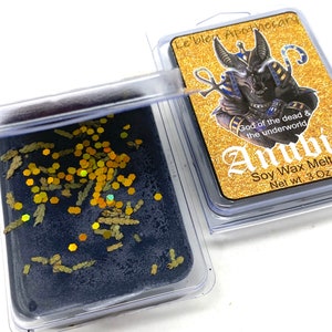 May include: A black soy wax melt with gold glitter and dried herbs in a clear plastic container. The label reads "Le bleu Apothecary", "God of the dead & the underworld", "Anubi", "Soy Wax Melt", and "Net wt. 3 Oz".