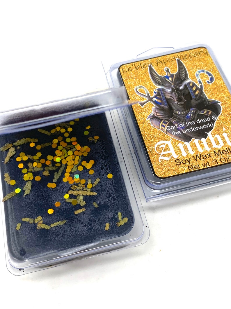 May include: A black soy wax melt with gold glitter and dried herbs in a clear plastic container. The label reads "Le bleu Apothecary", "God of the dead & the underworld", "Anubi", "Soy Wax Melt", and "Net wt. 3 Oz".